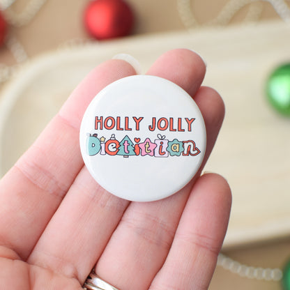 Button badge with 'Holly Jolly Dietitian' text held in a hand, with Christmas decorations in the background.