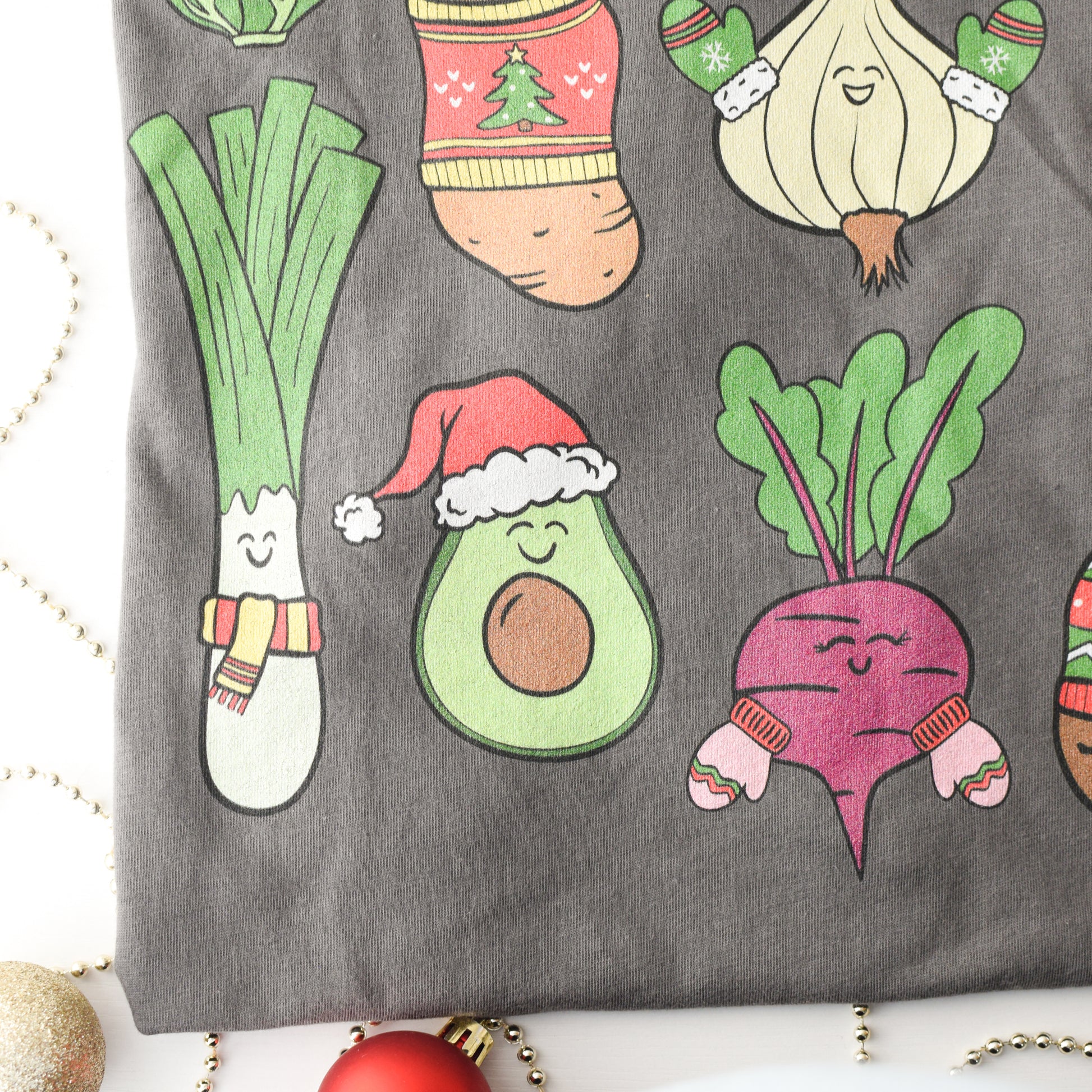 Close up of gray tshirt with cartoon avocado, beetroot, and leek wearing Christmas hats and scarves on a white background.