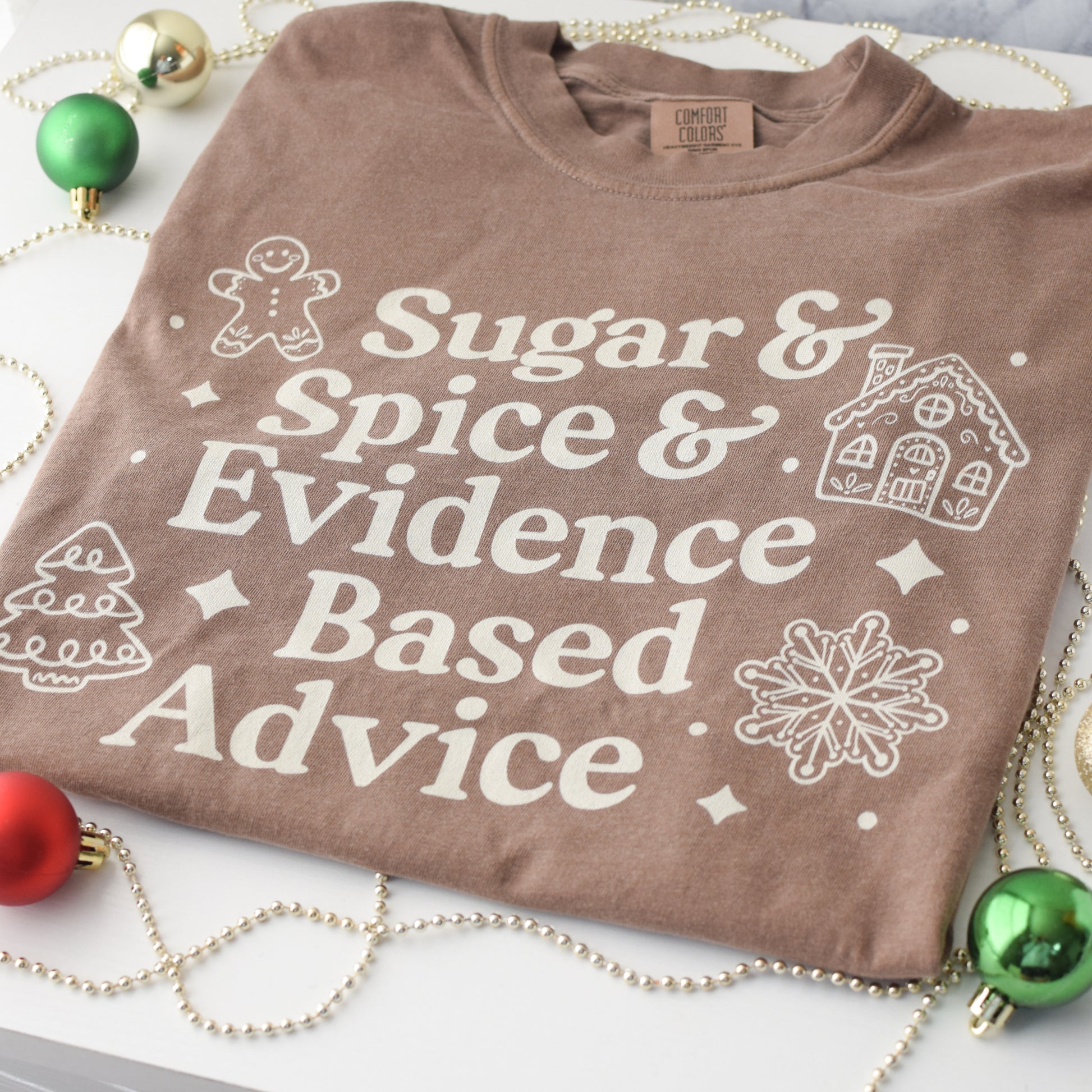 Brown t-shirt with white text and graphics on a white background with Christmas decorations.