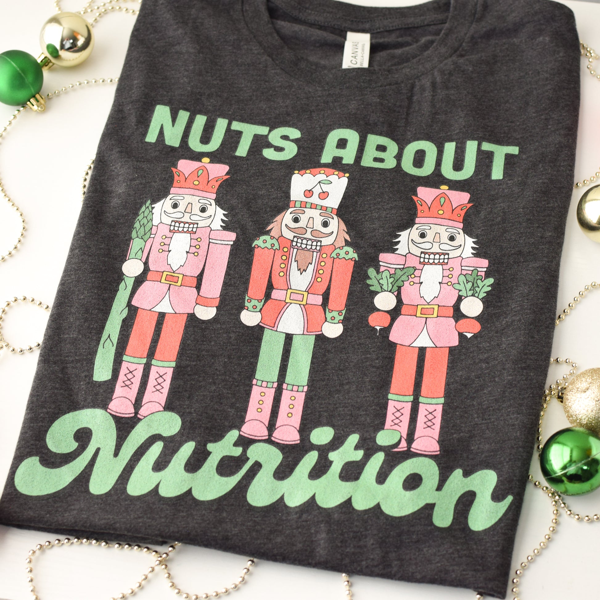 Dark gray t-shirt with nutcracker design and 'Nuts About Nutrition' text on a white background with Christmas decorations.