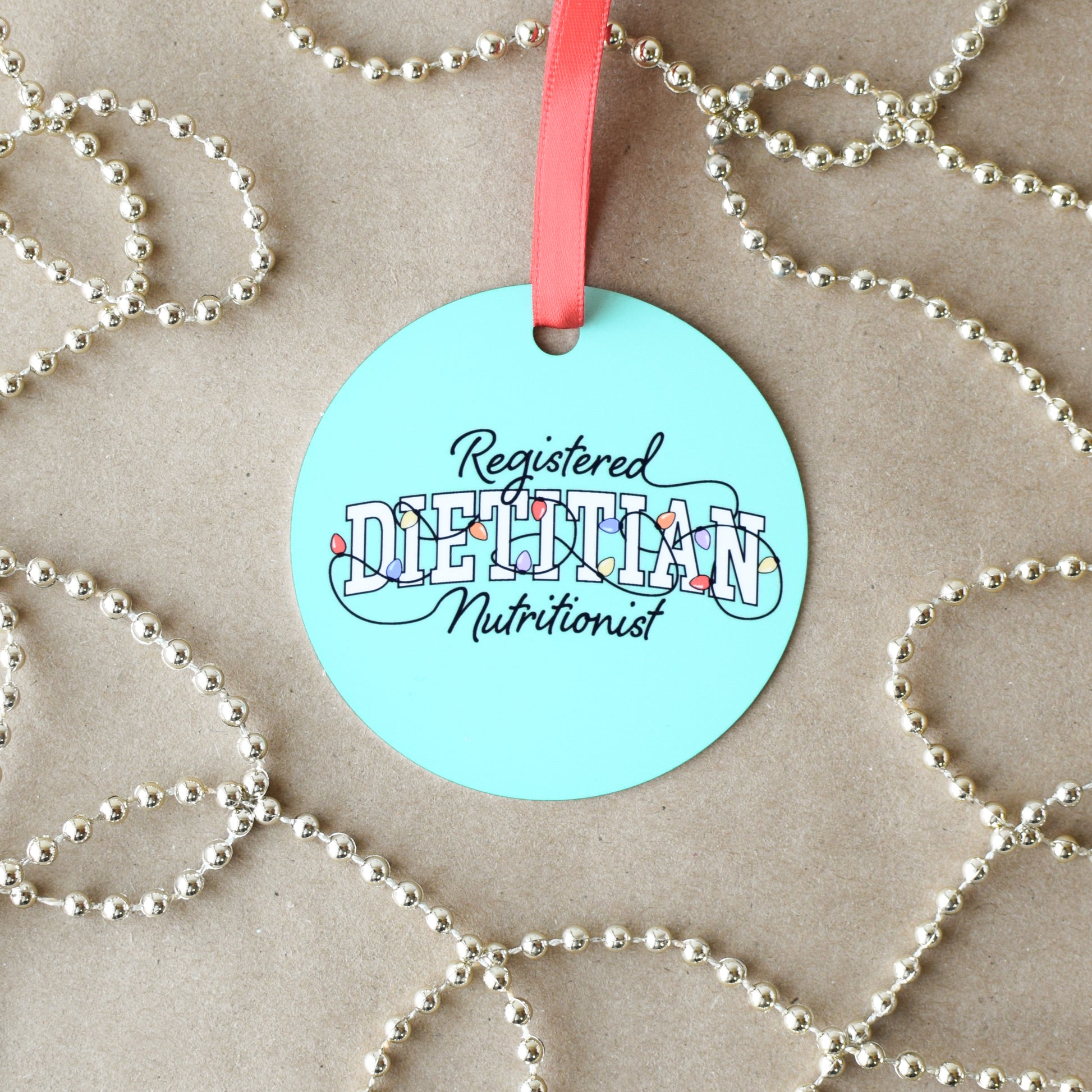 Round green-blue ornament with 'Registered Dietitian Nutritionist' text on a beige background with silver beads.
