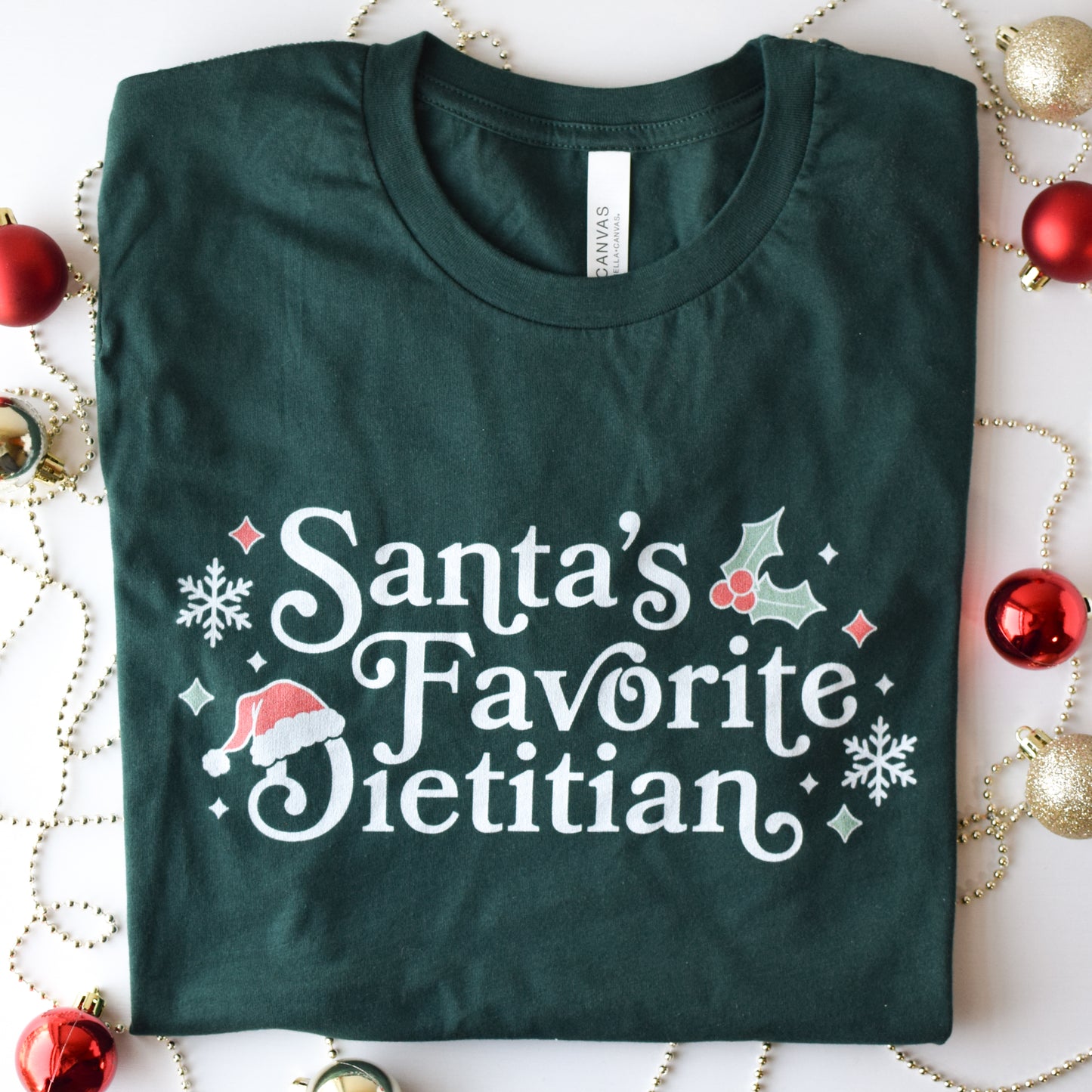 Folded green t-shirt with 'Santa's Favorite Dietitian' text on a decorative background with Christmas ornaments.