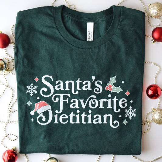 Folded green t-shirt with 'Santa's Favorite Dietitian' text on a decorative background with Christmas ornaments.