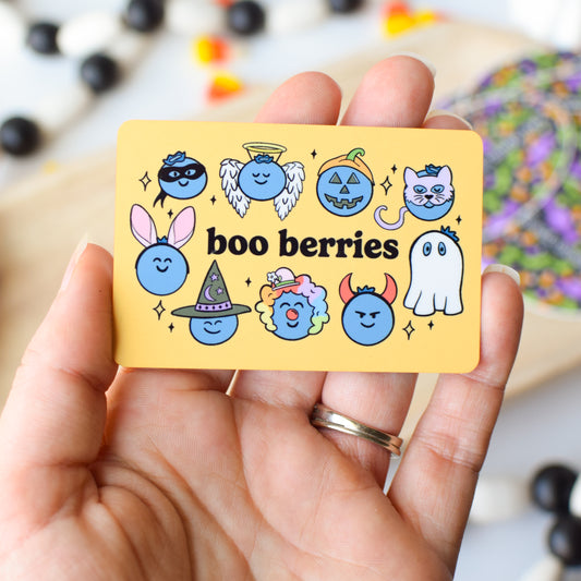 Hand holding a card with cartoon characters and 'boo berries' text, with Halloween-themed decorations in the background.