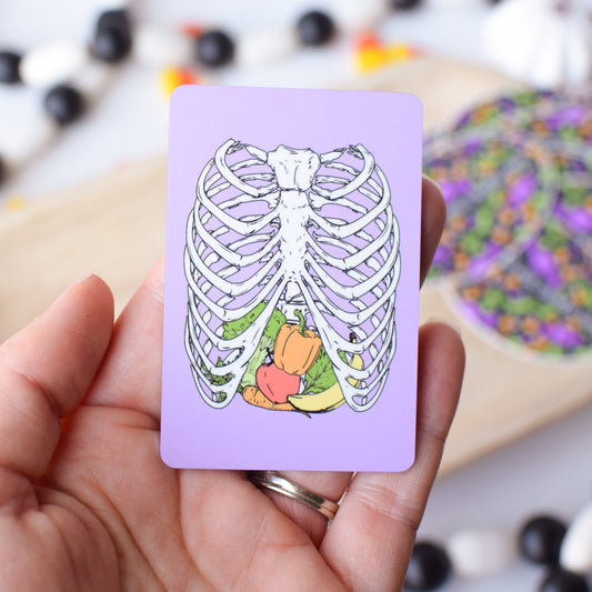 Hand holding a card with a skeleton design featuring fruits and vegetables on a blurred background.