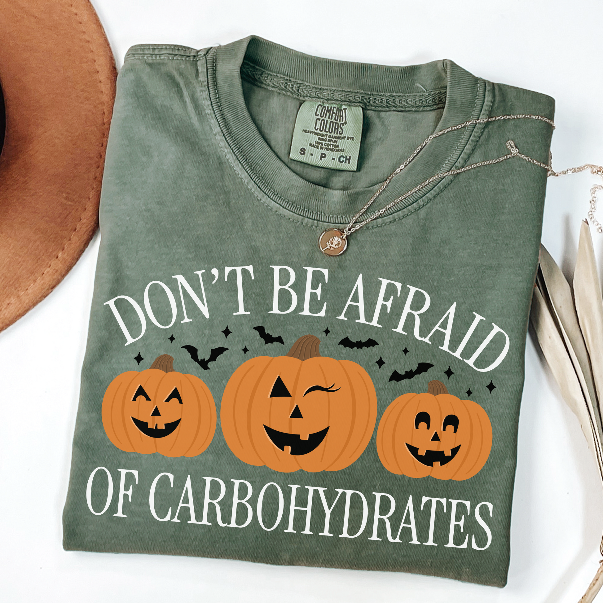 A green t-shirt with the text 'DON'T BE AFRAID OF CARBOHYDRATES' printed on the front, accompanied by images of three pumpkins. The shirt is presented with a hat and a necklace in the background, suggesting a casual, relaxed setting.